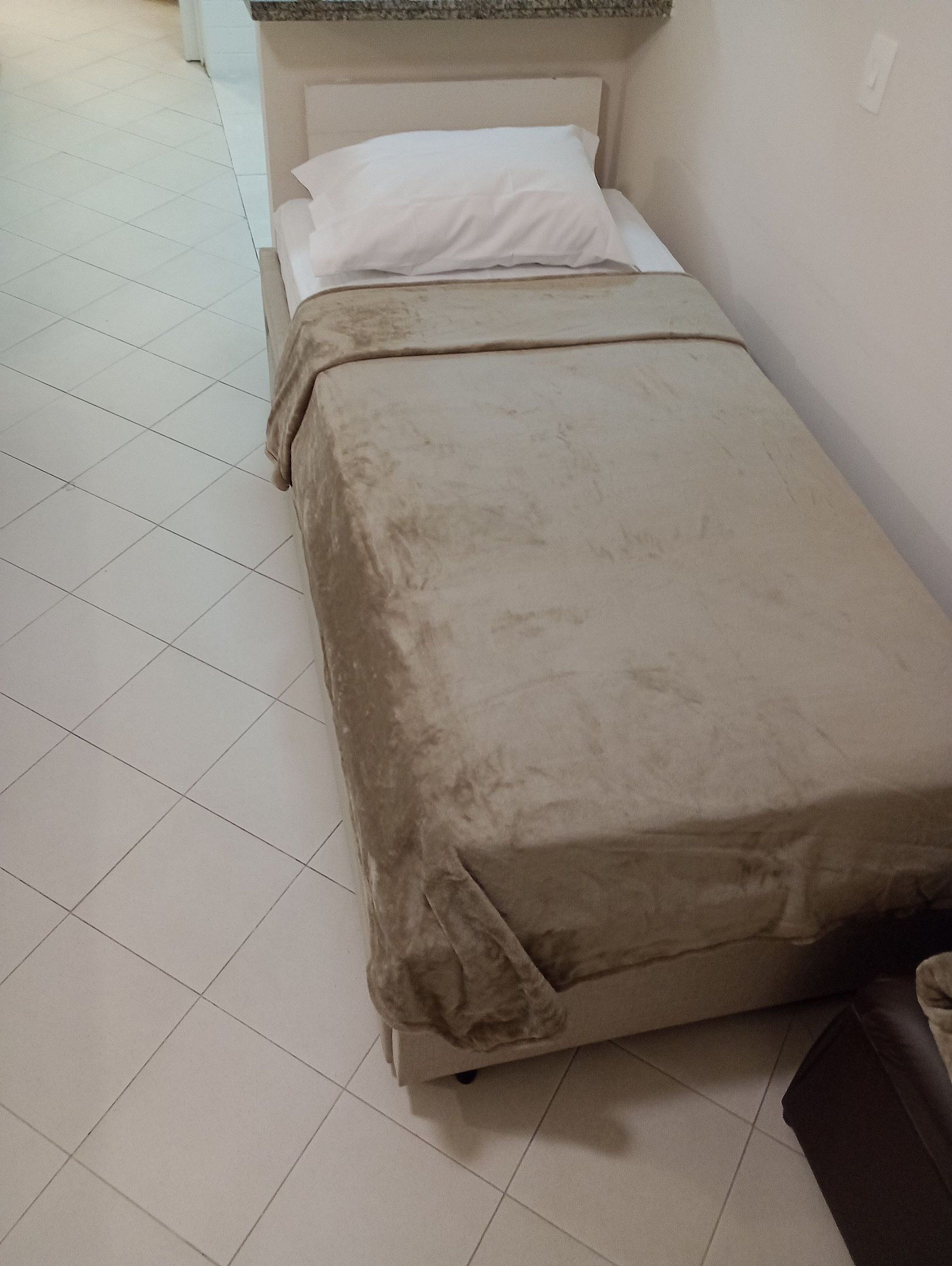 Ett litet rum med en enkel säng, ett vitt sänggavel, ett beige överkast och ett klinkergolv.