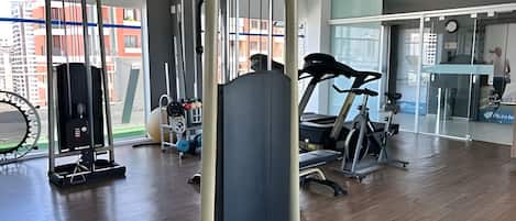 Un gimnasio con equipo de ejercicio, ventanas amplias y vistas a edificios.