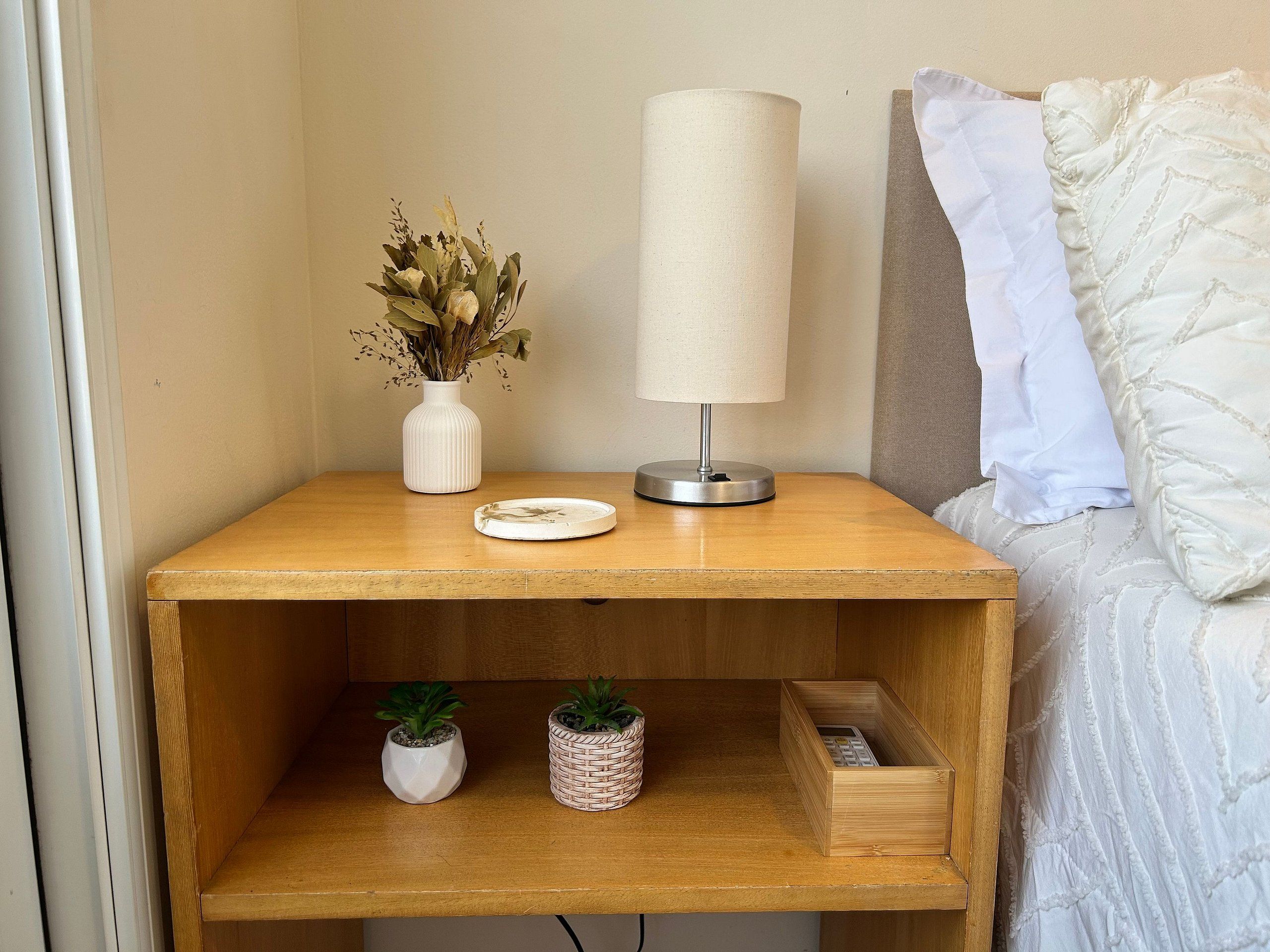 A wooden bedside table with a lamp, a vase with dried flowers, and two small potted plants.