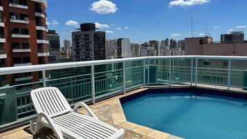 A rooftop pool with a clear view of the city skyline, a white lounge chair, and a glass railing.