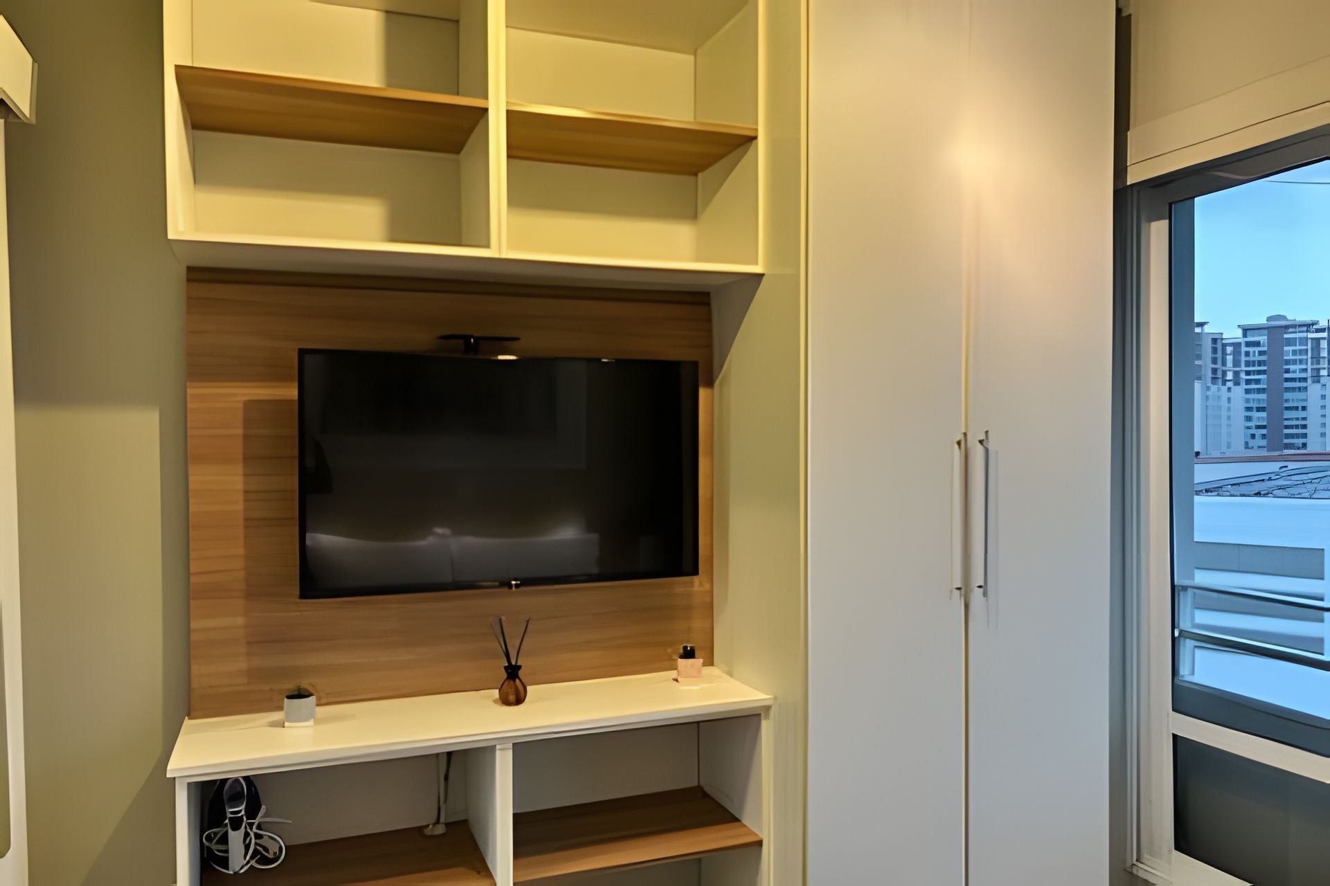 Ein modernes Zimmer mit einem an der Wand befestigten Fernseher, eingebauten Regalen und einem Fenster mit Blick auf Gebäude.