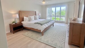 Un dormitorio con una cama grande, cabecera de madera, mesitas de noche con lámparas, una mesita auxiliar, cómoda y un ventanal con vistas a zonas verdes.
