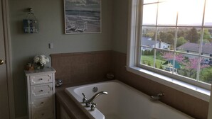 Une salle de bain avec une grande baignoire, une fenêtre donnant sur des maisons et des arbres, une commode blanche et un tableau encadré au mur.