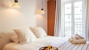 Hotelkamer met een bed, twee hanglampen, een raam met gordijnen en een ontbijttafel met croissants en sinaasappelsap.