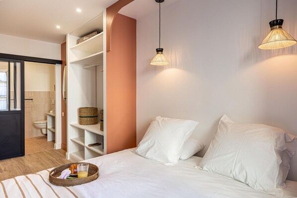Une chambre à coucher avec un lit, des oreillers blancs, une table de chevet avec un panier et un luminaire suspendu.