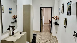 Un corridoio con pavimento in terrazzo, una parete bianca con quadri appesi, uno specchio e un piccolo vaso con fiori secchi.