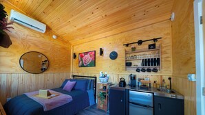 Une petite pièce comprenant un lit, un coin cuisine, un miroir, une horloge et une étagère avec des ustensiles de cuisine.