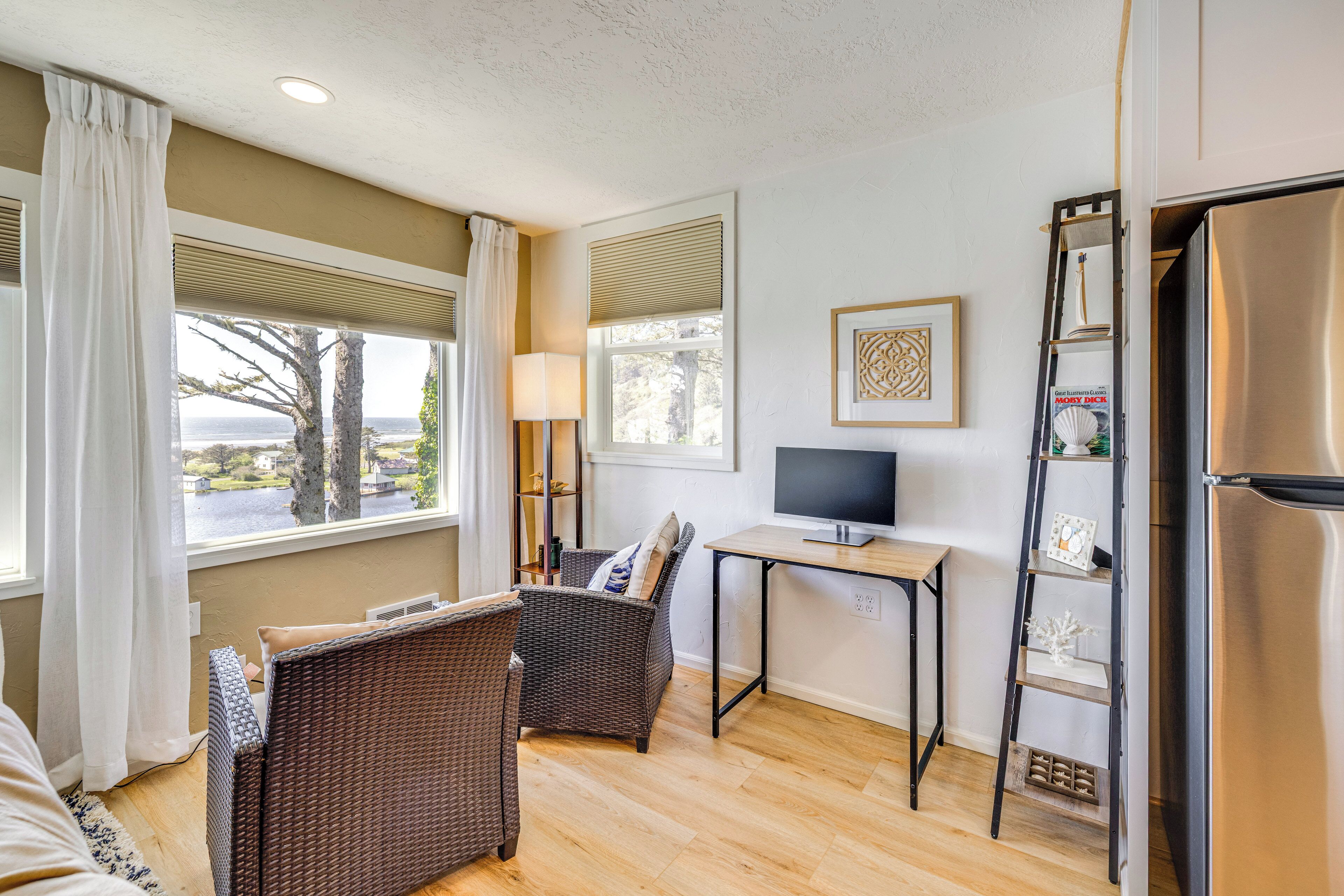 Una habitación con sofá, dos sillones de mimbre, una mesa pequeña con televisor, refrigerador y ventana con vista a árboles y agua.