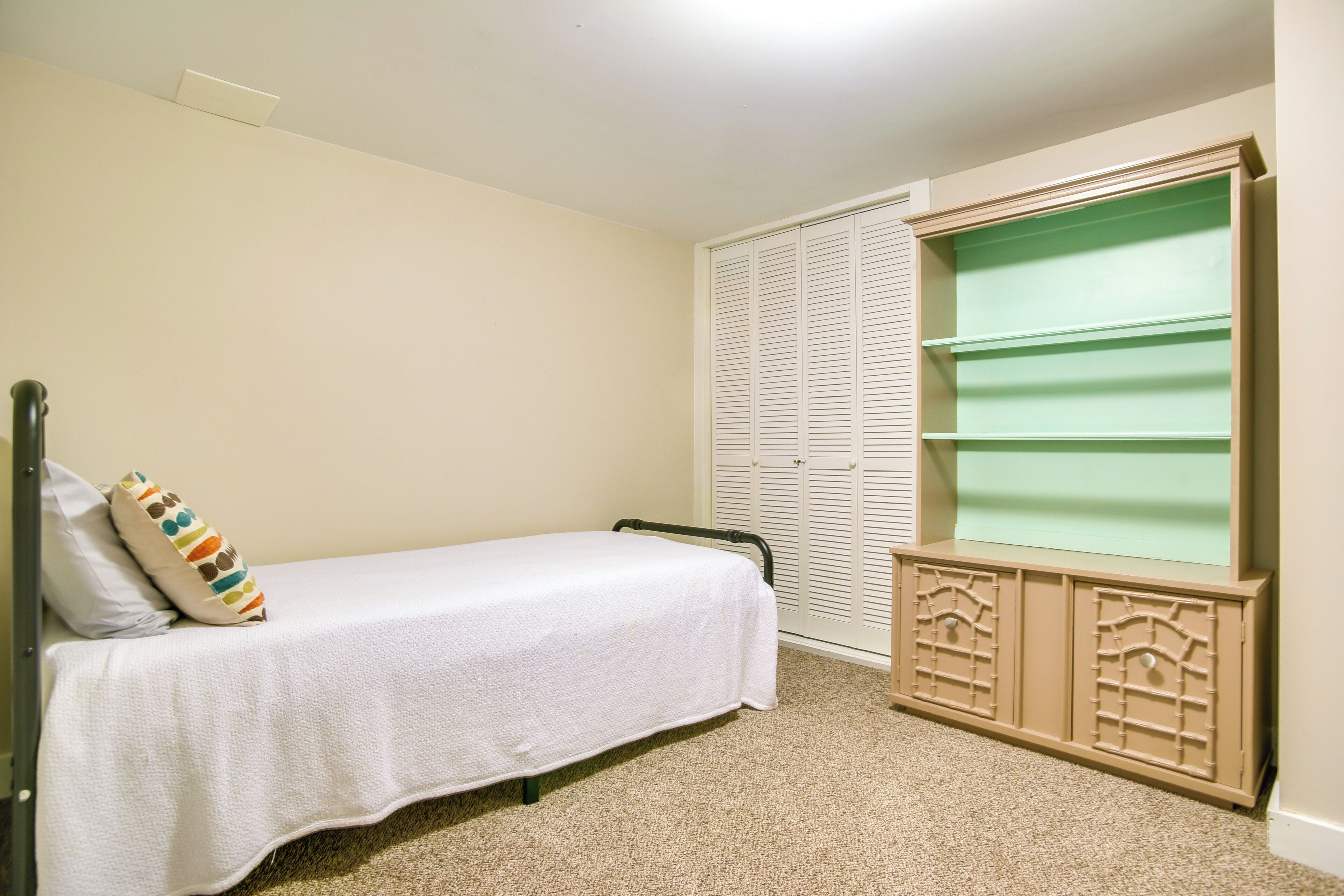 Une chambre avec un lit simple, une armoire dotée d’un miroir vert et un sol recouvert d’une moquette.