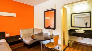 A hotel room with a single bed, a small table with a chair, a mirror, and a bathroom with a sink and mirror. - Economy Hotel Kansas City (Blue Springs)