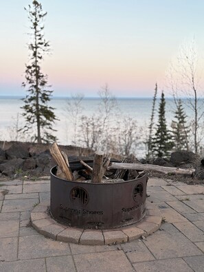 A metal fire pit with the inscription "Superior Shores" on a paved area near a body of water with trees in the background. - Sunrise Suite on Lake Superior- Pool & Hot Tub (Two Harbors)
