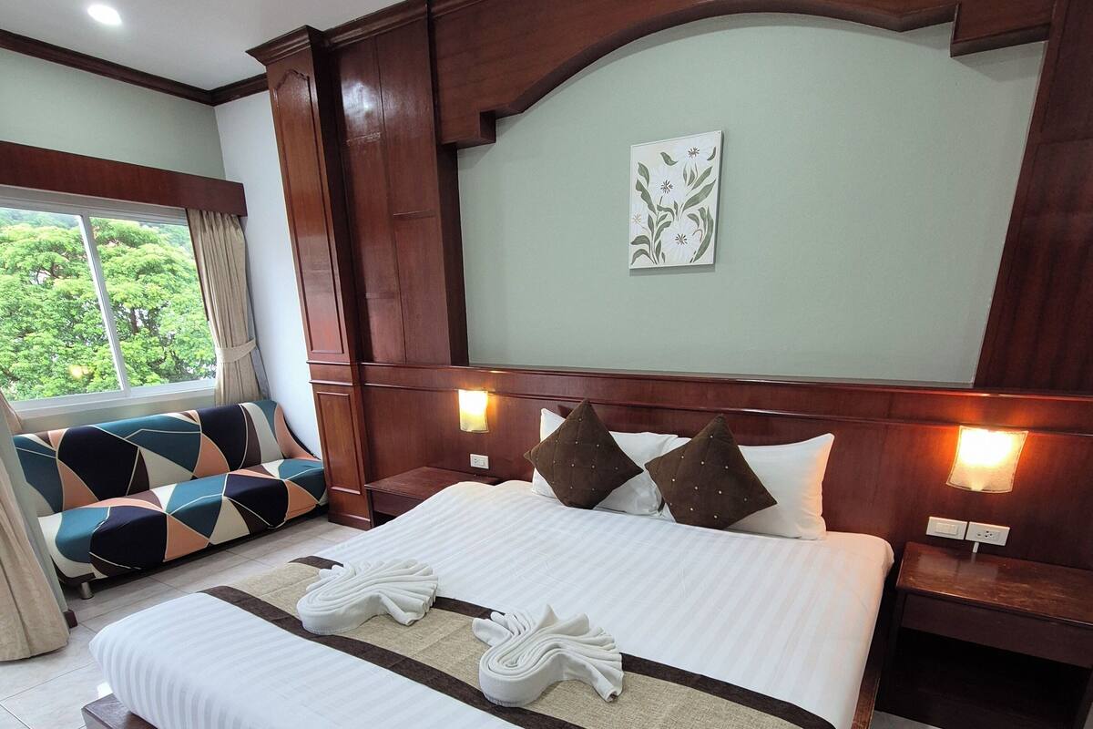 A hotel room with a large bed, two bedside tables, a window with a view of greenery, and a wall-mounted artwork.