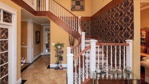 Eine imposante Treppe mit hölzernem Geländer und weißen Spindeln, die zu einer Empfangshalle mit ornamentalem Wandpapier und einer Wandleuchte führt.