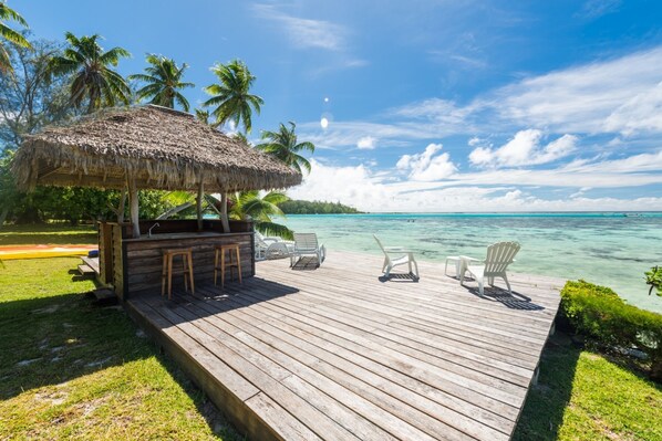 A wooden deck with lounge chairs and a thatched-roof structure, overlooking a turquoise sea and palm trees.