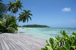 A wooden deck leading to a turquoise sea with palm trees, boats, and a forested island in the background.