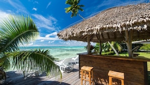 Een houten terras met rieten dak, witte lounge stoelen en een houten bar, met uitzicht op een heldere blauwe zee.