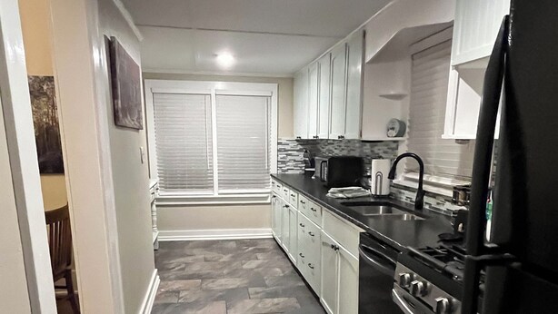 A kitchen with a stone floor, white cabinets, a black refrigerator, and a window with blinds.