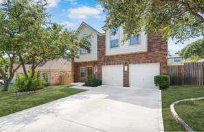 Two-story home with a brick facade and a gabled roof, featuring a front driveway and a small yard with trees.