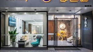 A hotel entrance with a modern design, featuring a reception area with a glass facade, a seating area with a sofa and chairs, and a decorative wall with a circular artwork.
