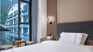 A hotel room with a large window offering a view of a mountain and buildings. There is a bed with white linens, a bedside table with a lamp, and a wall-mounted light fixture.