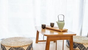 A wooden table with a woven mat, two woven stools, and a ceramic pitcher on the table.