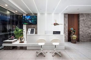 Uma área de recepção com balcão, duas cadeiras, uma tela de TV e uma planta decorativa.