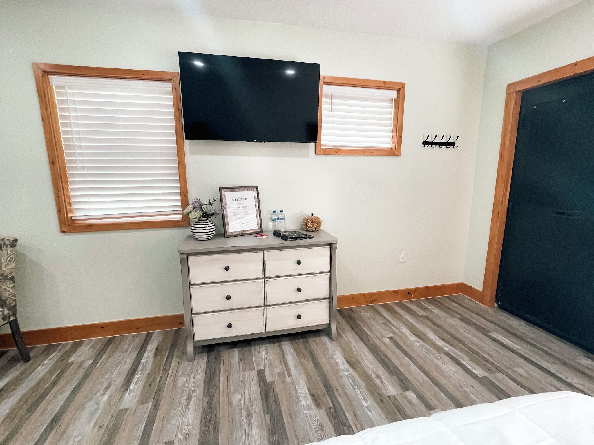 A bedroom with a bed, a dresser with drawers, a television mounted on the wall, and two windows with blinds.