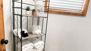 Une étagère en métal avec des serviettes pliées, une petite plante en pot et un pichet blanc sur lequel est inscrit le mot « Béni ».