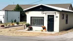 Une maison de plain-pied avec une porte noire et des volets aux fenêtres, un petit porche agrémenté de plantes en pot et un ciel d’un bleu limpide.