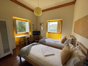 Un dormitorio con dos camas, un escritorio, un televisor y ventanas que dan a vistas de vegetación y un edificio.