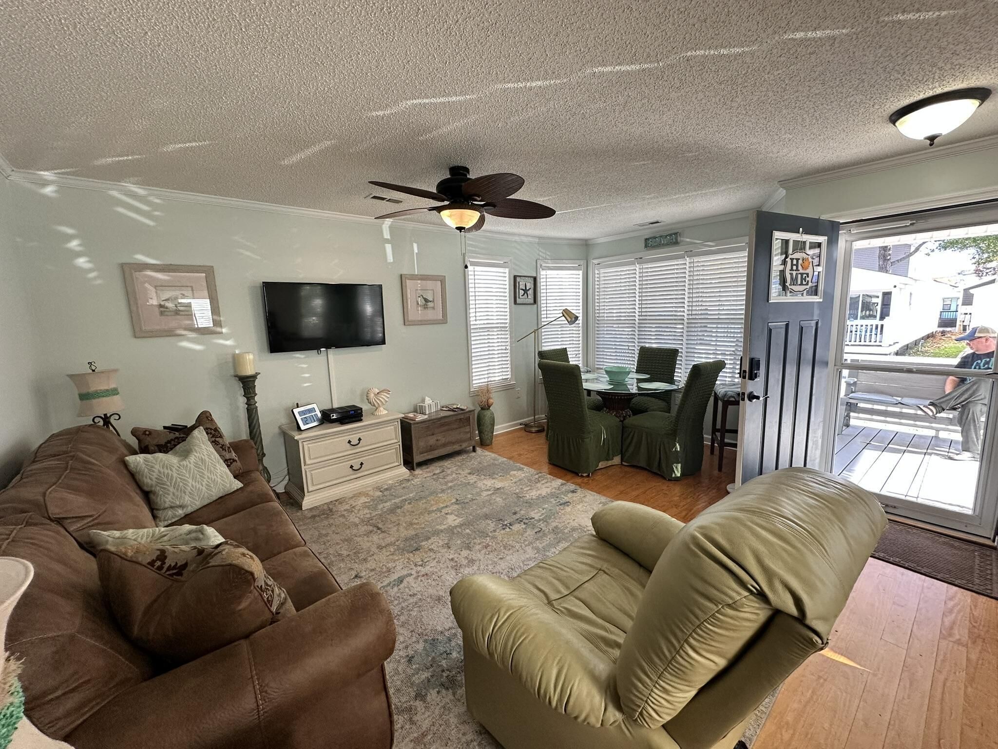 Ein Wohnzimmer mit einem braunen Sofa, einem grünen Sessel, einem Fernseher, einem Deckenventilator und einem Essbereich mit Tisch und Stühlen.