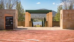 Ein Steinweg führt zu einem Parkeingang mit einer Pergola über dem Wasser, umgeben von Bäumen und Hügeln.