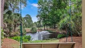 A swing chair overlooking a pond with a fountain, surrounded by lush greenery and trees. - 2889 - Two-Story Villa | Golf Views | Sea Pines (Hilton Head Island)