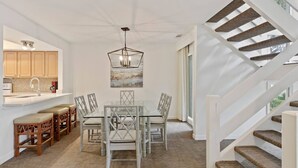 A spacious dining area with a glass table and chairs, a modern chandelier, and a staircase with wooden steps. - 2889 - Two-Story Villa | Golf Views | Sea Pines (Hilton Head Island)
