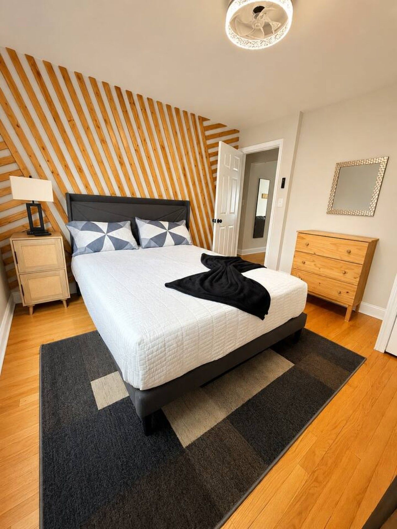 A bedroom with a bed, a nightstand, a dresser, a mirror, and a wall with a striped pattern.
