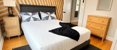 A bedroom with a bed, a nightstand, a dresser, a mirror, and a wall with a striped pattern.