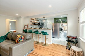 A modern living room with a sofa, a coffee table, and a kitchen area with a refrigerator, microwave, and bar stools.