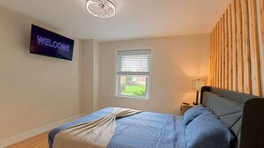 Quarto com cama, televisão exibindo a mensagem 'WELCOME', janela e painel de parede em madeira.