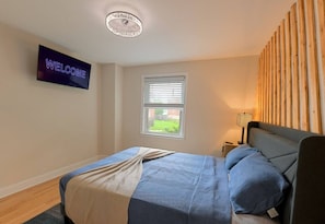 Une chambre à coucher avec un lit, un téléviseur affichant le mot « BIENVENUE », une fenêtre avec des stores et une lampe sur une table de chevet.