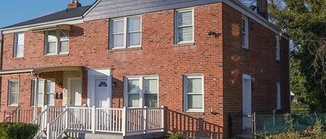 A two-story brick house with a front porch, a small front yard, and a clear sky.