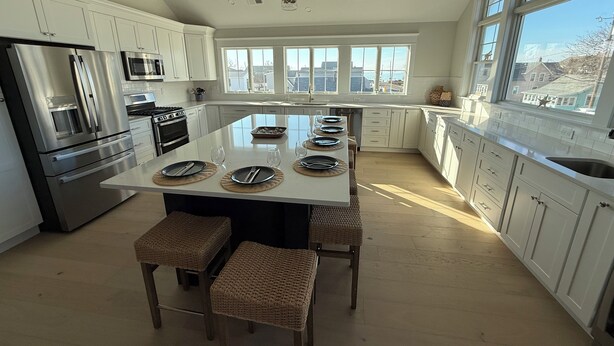 A modern kitchen with white cabinets, stainless steel appliances, a large island with seating, and large windows offering views of the outside. - “The Delford” Beach House only Steps to the Bay Beaches (Lower Township)
