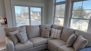 A beige sectional sofa with patterned cushions, a woven rug, and a view of a residential street through large windows. - “The Delford” Beach House only Steps to the Bay Beaches (Lower Township)