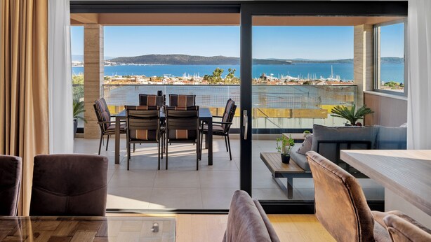 A modern living room with a glass door leading to a balcony overlooking a coastal area with boats and buildings.