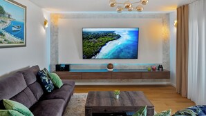 A modern living room with a large flat-screen TV mounted on the wall, a dark brown sofa with green cushions, a wooden coffee table, and a marble accent wall.