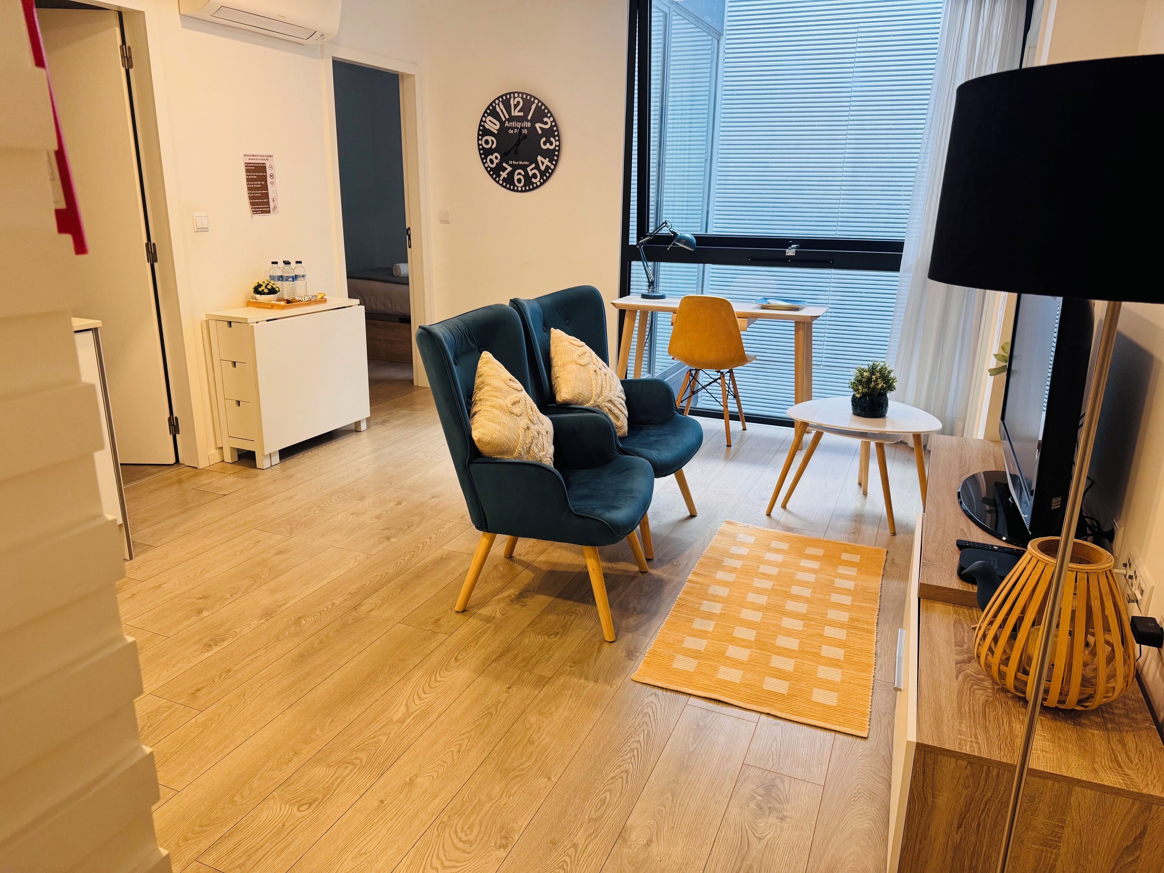 A modern living room with a dark blue sofa, a small round table, and a TV. There is a wooden floor and a large window with blinds.