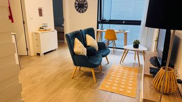 A modern living room with a dark blue sofa, a small round table, and a TV. There is a wooden floor and a large window with blinds.