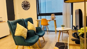 A modern living room with a teal sofa, wooden flooring, a round table, and a large window with blinds.