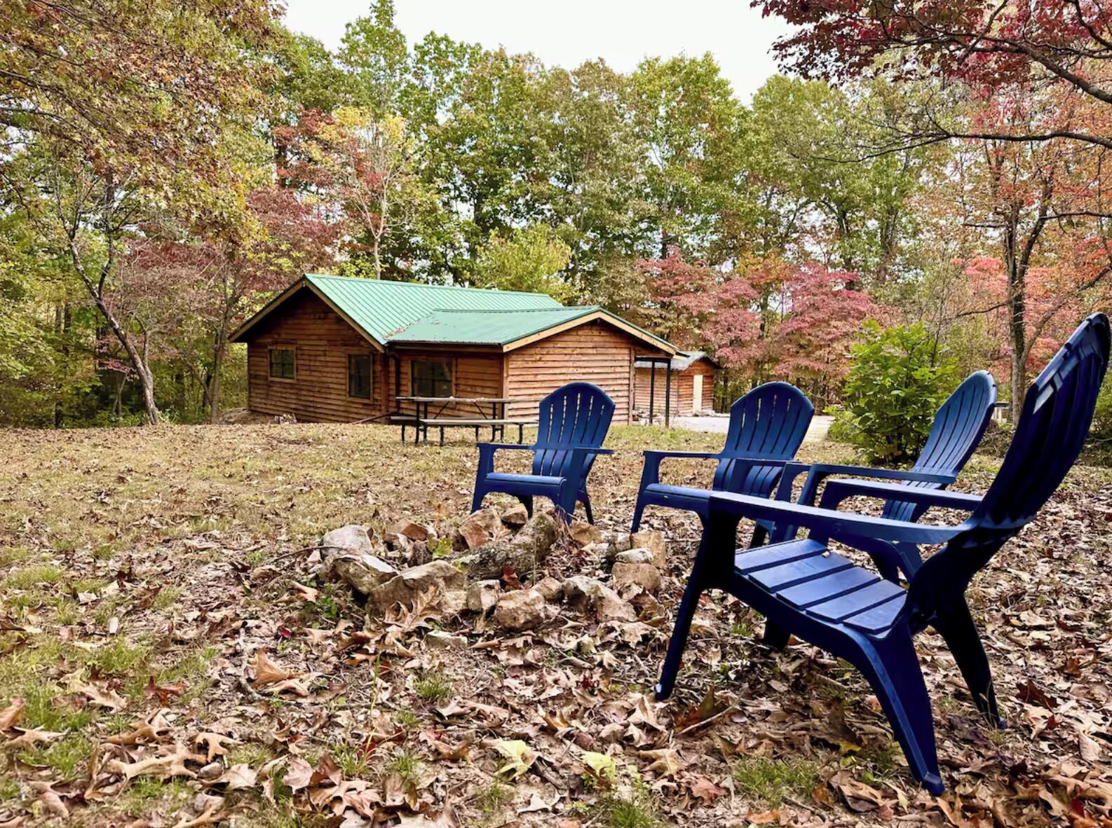 Een rustieke blokhut met een groen dak, omringd door bomen met herfstbladeren. Drie blauwe Adirondack-stoelen en een picknicktafel bevinden zich voor de blokhut.