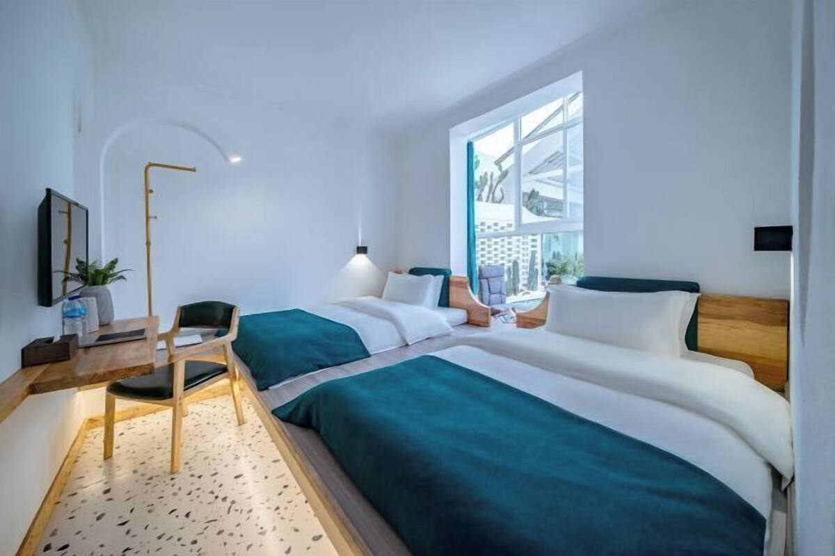 A modern hotel room with two beds, a desk, a chair, a lamp, and a large window.