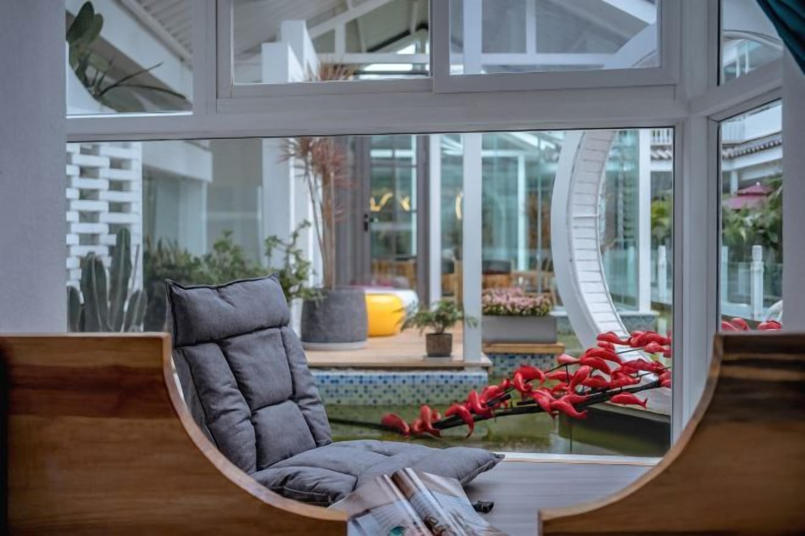 Un intérieur moderne avec une fenêtre incurvée, un fauteuil rembourré de couleur grise et une vue sur un espace extérieur agrémenté de plantes et d’un élément décoratif aquatique.